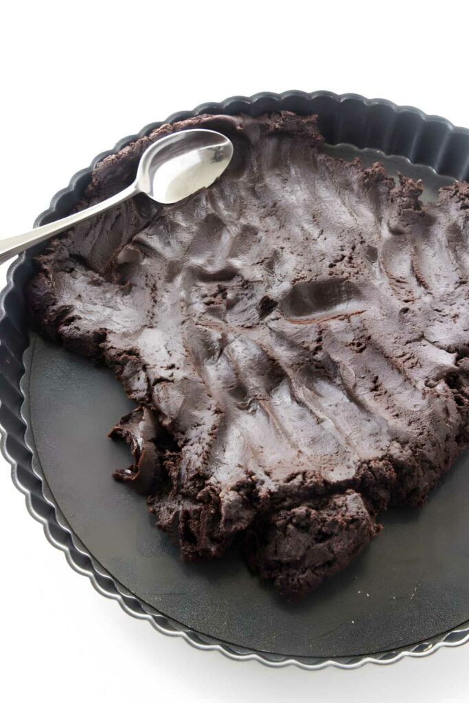 Spoon pressing chocolate dough into tart pan on white background.
