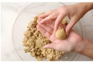Hands shaping dough ball over bowl with crumbly mixture on white surface.