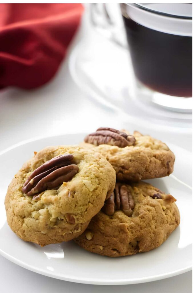 Stacked pecan cookies on a white plate, blurred coffee and red cloth behind.