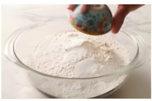Hand holds small bowl over glass mixing bowl with flour on white surface.