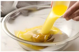 Hand pours melted butter into a clear glass mixing bowl on white surface.