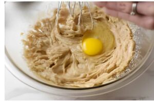 Hand whisking dough with cracked egg in glass bowl.