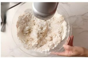 Hand using electric mixer blends flour in glass bowl on white countertop.