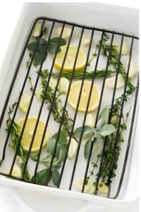 White roasting pan with rack, lemon slices, garlic cloves, and fresh herbs underneath.