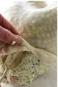 Hand spreading herb butter under raw turkey skin with a spoon.