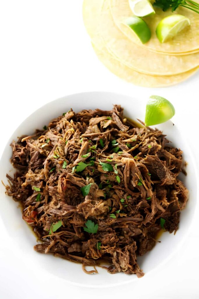 Bowl of shredded beef with herbs and lime, tortillas in background.