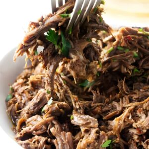 Shredded beef pulled apart with forks in a white bowl, topped with herbs.