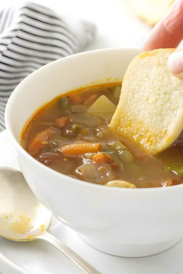Hand dipping bread into Italian vegetable soup; spoon and striped cloth beside bowl.