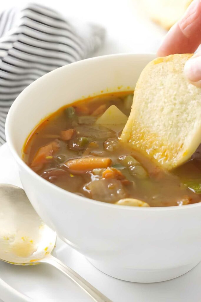 Hand dipping bread into Italian vegetable soup; spoon and striped cloth beside bowl.