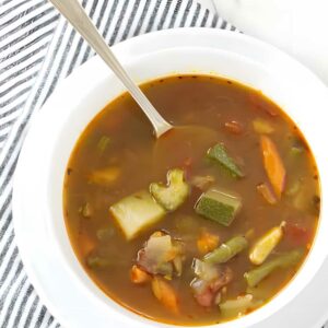 White bowl of chunky vegetable soup with silver spoon on striped cloth.