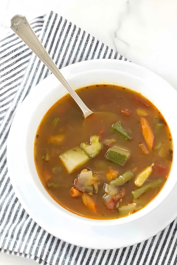White bowl of chunky vegetable soup with silver spoon on striped cloth.