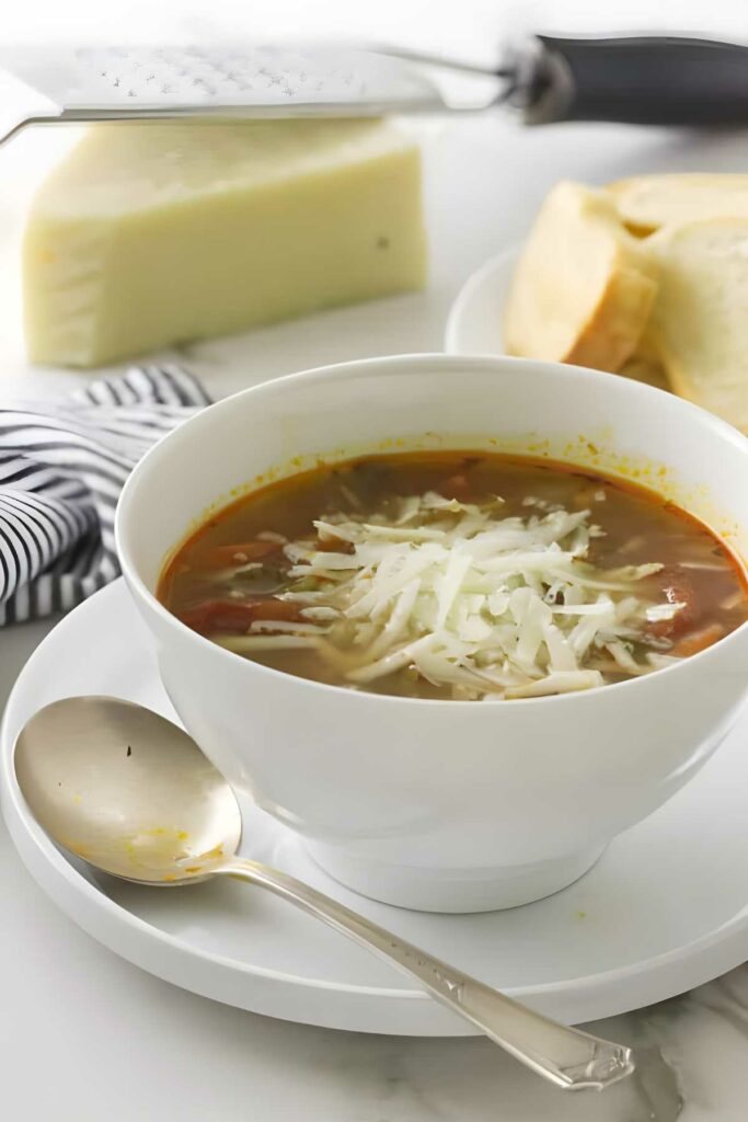 Bowl of minestrone soup with shredded cheese, served on a plate with spoon.