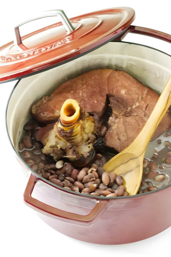 Red Dutch oven with beans, ham hock, and spoon inside.