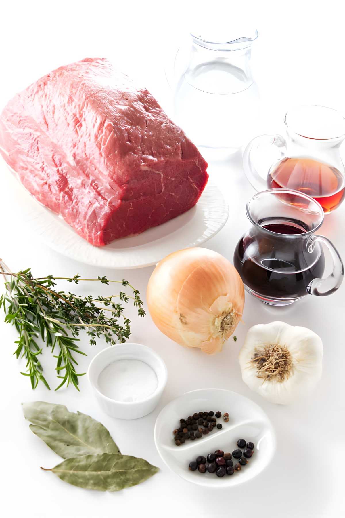Raw beef roast on plate with herbs, onion, garlic, seasonings, wine, water.