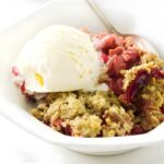 Strawberry rhubarb crisp with vanilla ice cream in a bowl, spoon on side.