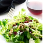 Green salad with walnuts, cranberries, gorgonzola cheese on white plate; wine behind.