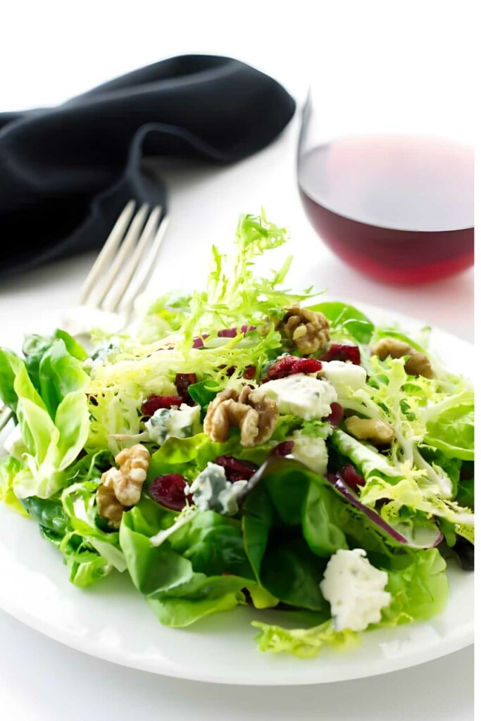 Green salad with walnuts, cranberries, gorgonzola cheese on white plate; wine behind.