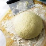 Ball of pizza dough on floured wood with dough scraper and flour bowl nearby.