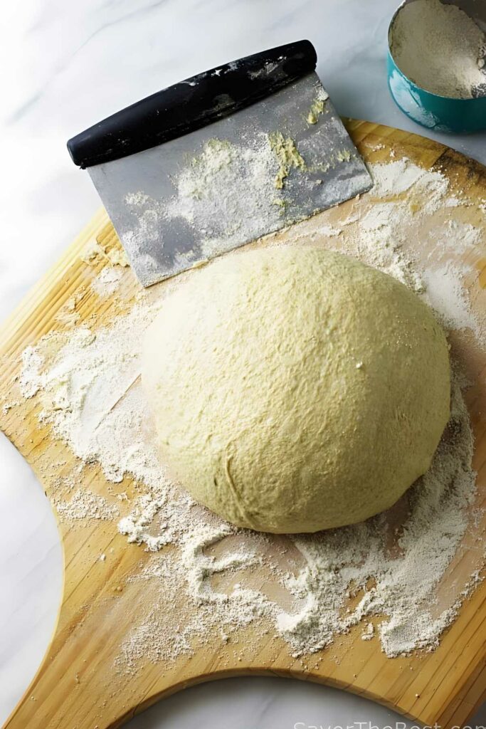 Ball of pizza dough on floured wood with dough scraper and flour bowl nearby.