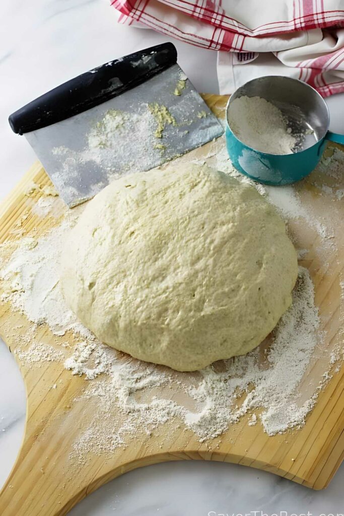 Einkorn pizza dough on a floured surface with scraper, measuring cup, and towel nearby.