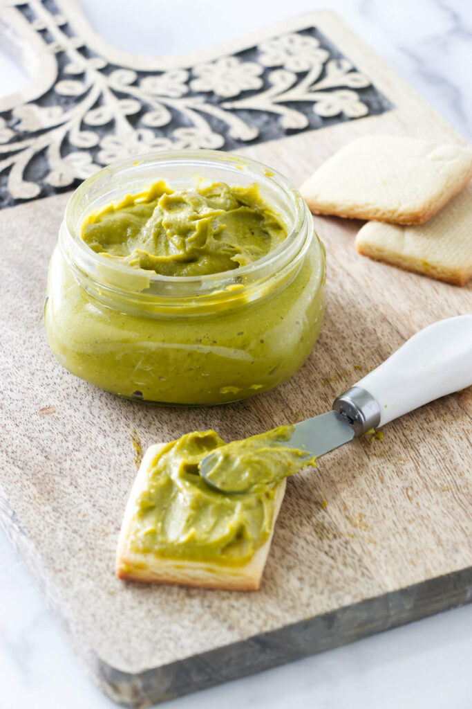 A jar of fresh pistachio paste in front of a cracker with pistachio butter.