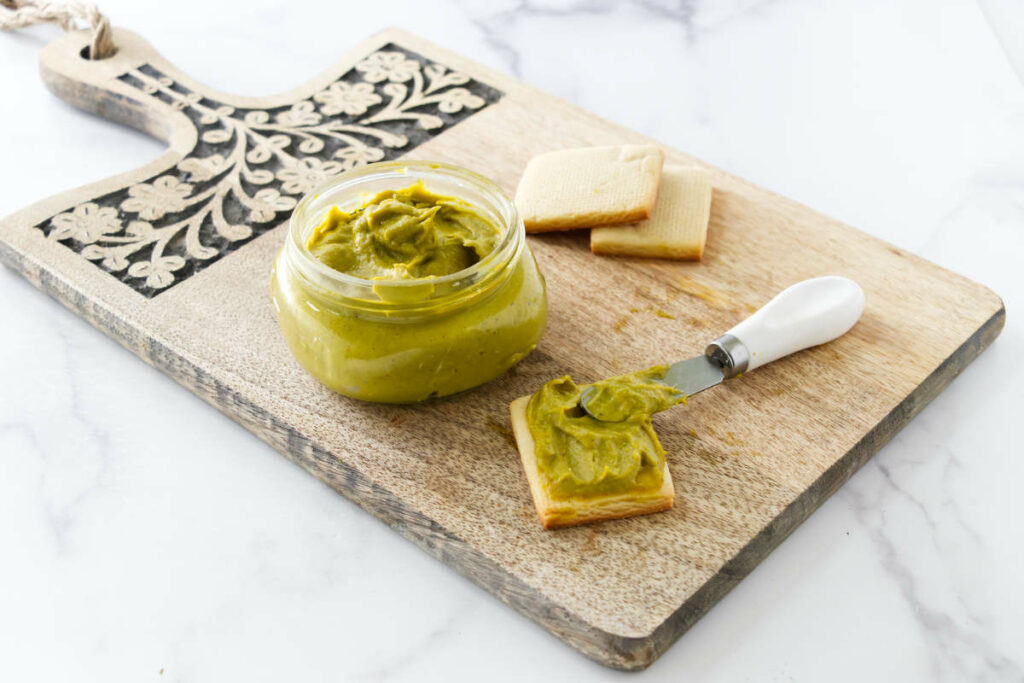 Spreading pistachio butter on a cracker next to a jar of pistachio paste.