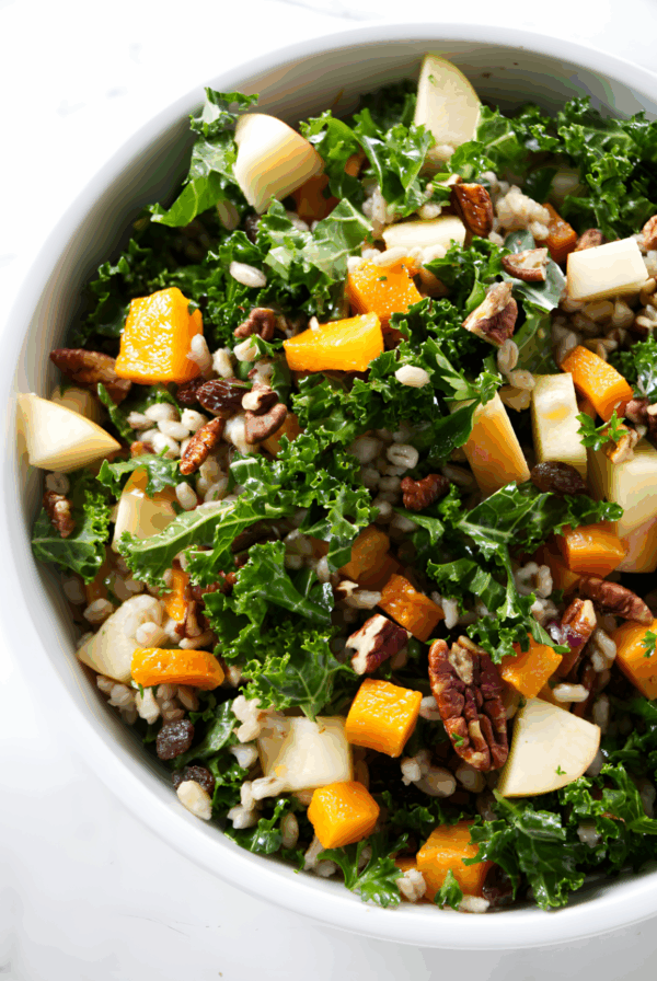 Colorful kale salad with squash, apples, pecans, barley, and raisins in bowl.
