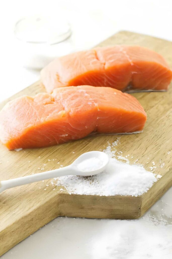 Two raw salmon fillets on a wooden board with salt and spoon nearby.