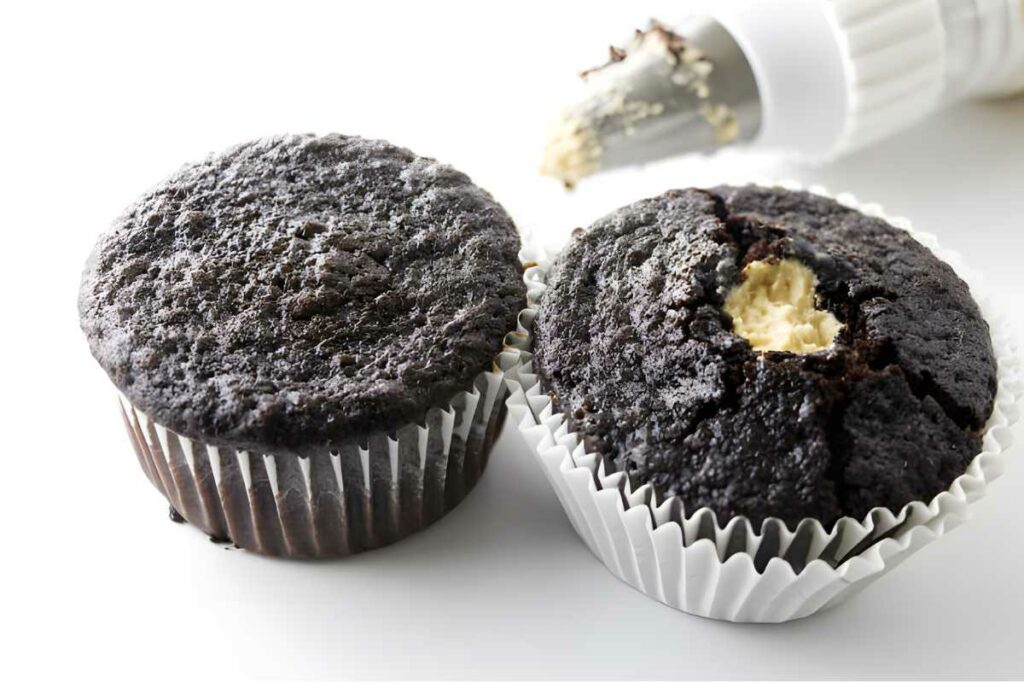 Two chocolate cupcakes, one with visible cream filling; piping bag in background.