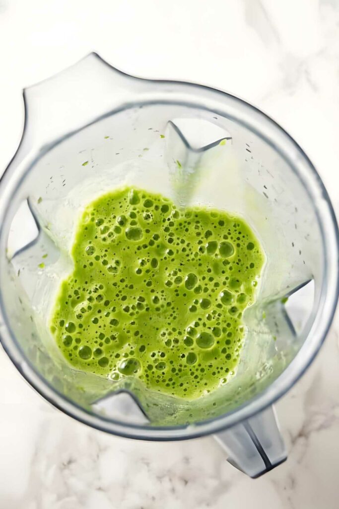 Blender with frothy green smoothie, viewed from above on white marble.