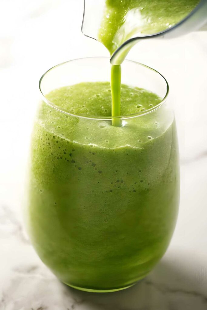 Green smoothie poured from blender into glass on white marble.