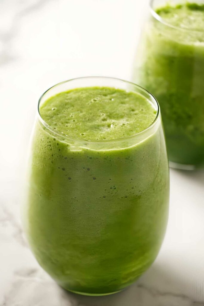 Two clear glasses of thick green smoothie on a light surface.
