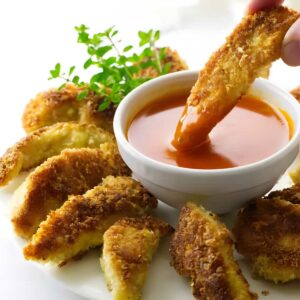 Hand dips golden-brown fried slice into orange sauce, with herbs on plate.