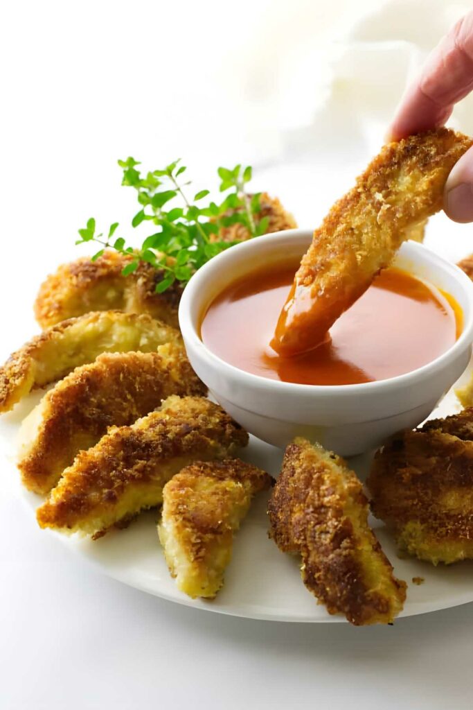 Hand dips golden-brown fried slice into orange sauce, with herbs on plate.