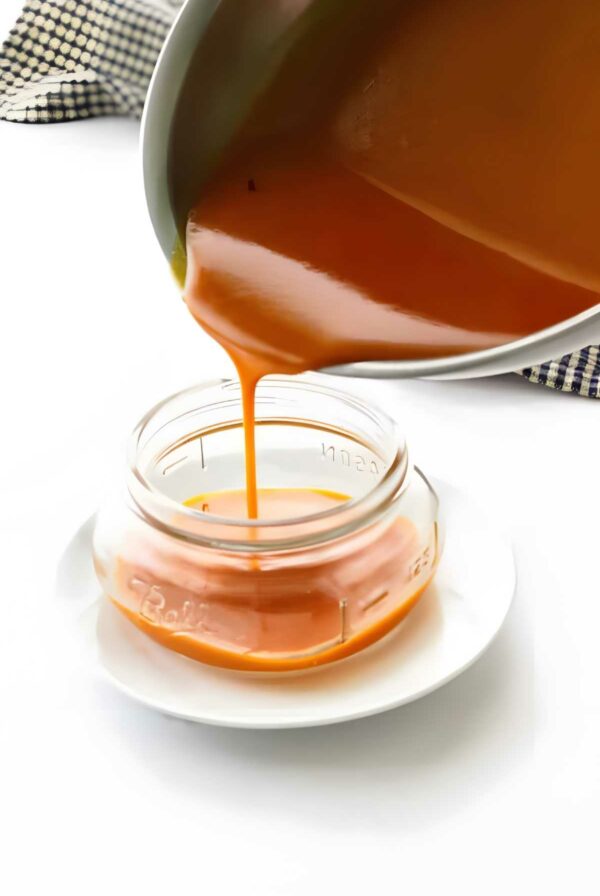 Caramel sauce poured from saucepan into mason jar on plate, checkered cloth behind.