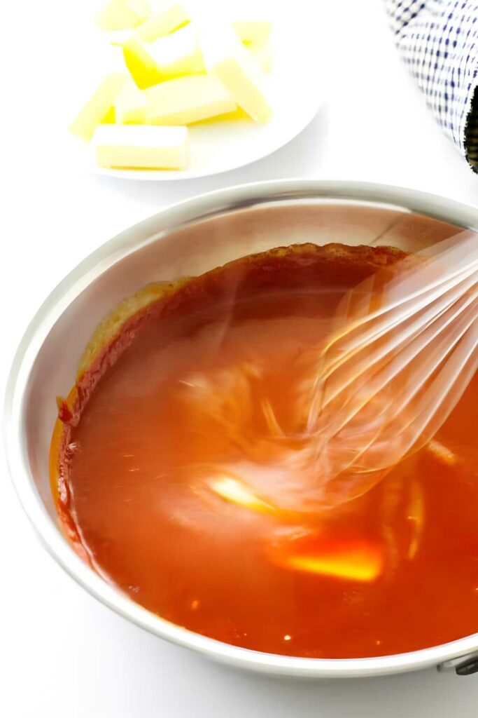 Red-orange sauce being whisked in saucepan with melting butter pat.