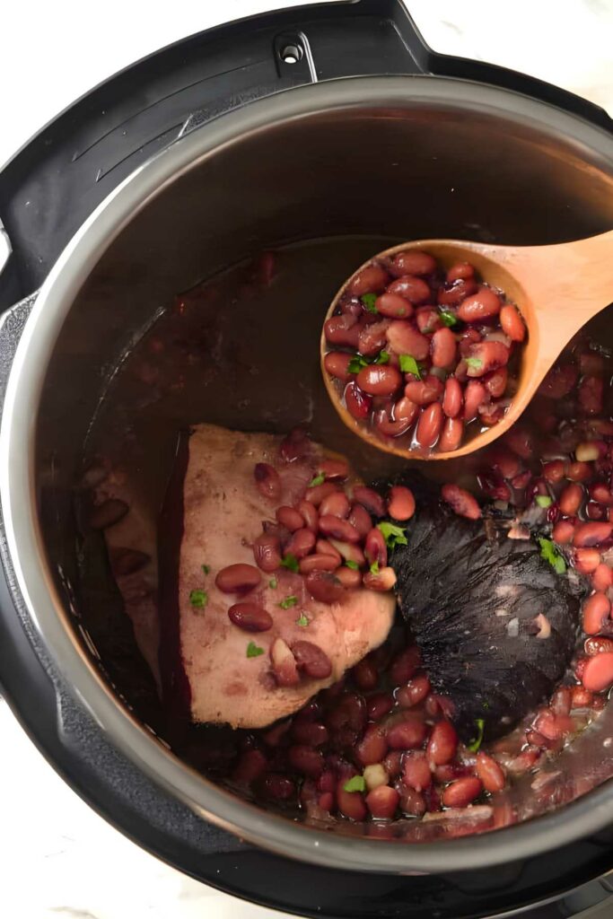 Wooden spoon lifts red beans and broth from Instant Pot with meat.
