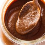 Spoon scooping creamy chocolate hazelnut spread from a glass jar.