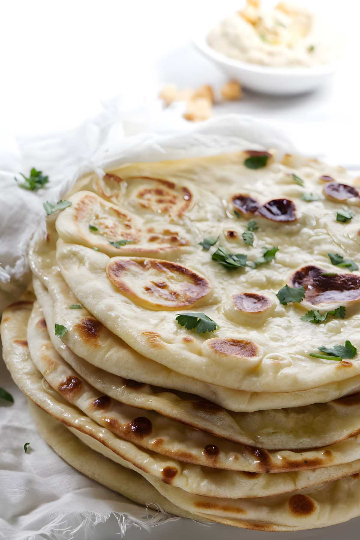 Stack of naan bread with cilantro on white cloth, dip blurred behind.