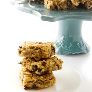 Three oat and raisin bars stacked on a white plate, more on blue stand.