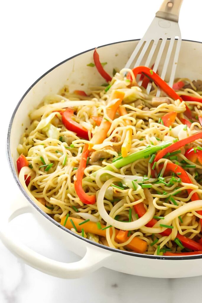 Stir-fried noodles with colorful vegetables in a white pan, spatula mixing.