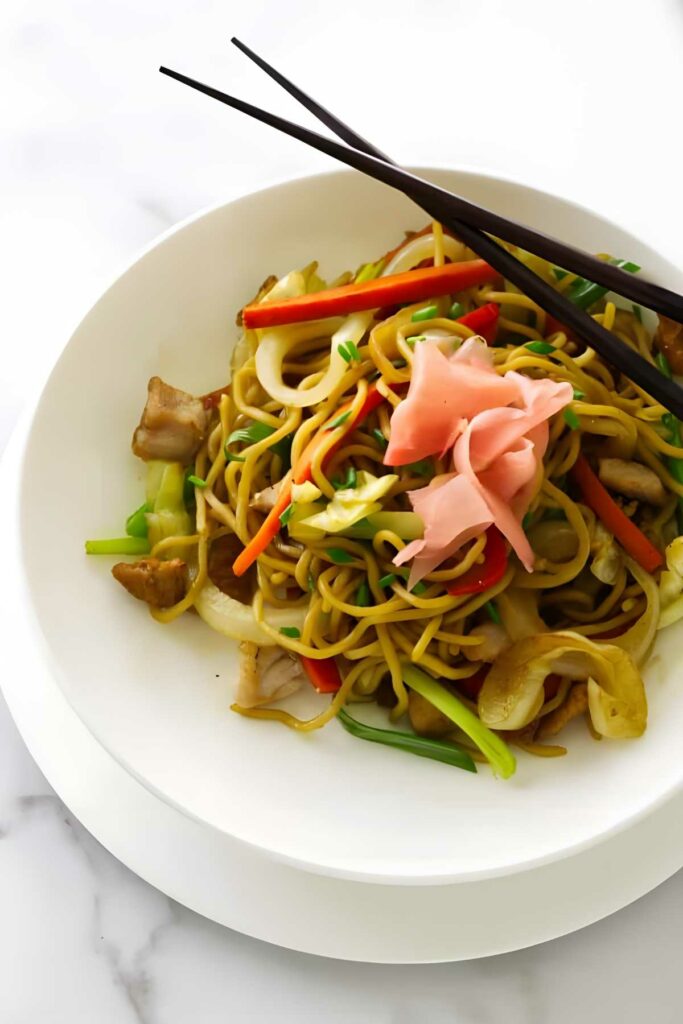 Stir-fried noodles with vegetables and meat on a white plate with chopsticks.
