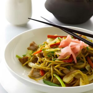 Stir-fried noodles with vegetables and chopsticks on plate; teapot and cup behind.
