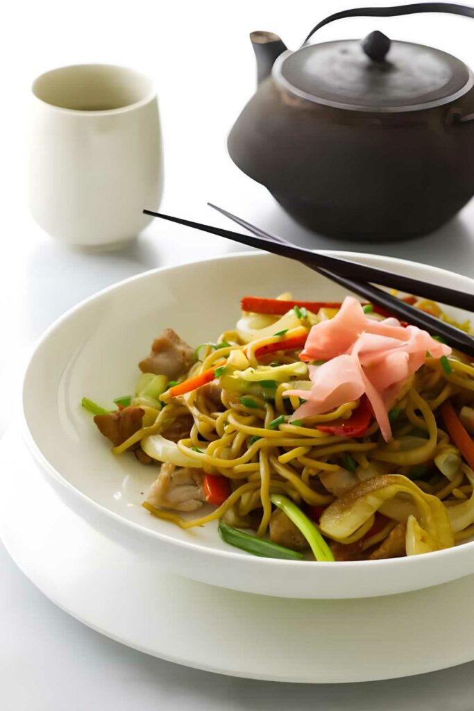 Stir-fried noodles with vegetables and chopsticks on plate; teapot and cup behind.