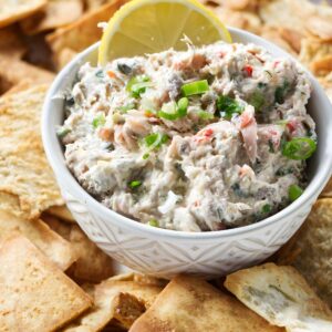 Bowl of creamy salmon spread with green onions, lemon slice, and pita chips.