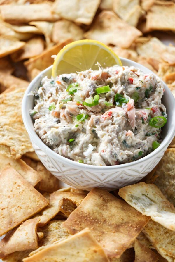 Bowl of creamy salmon spread with green onions, lemon slice, and pita chips.