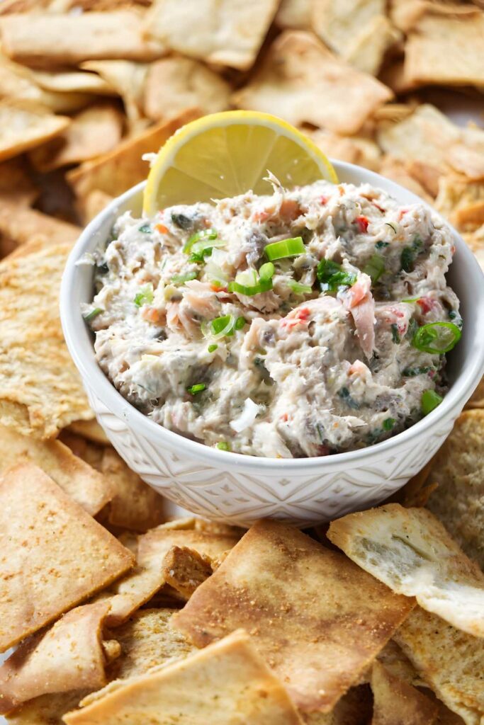 Bowl of creamy salmon spread with green onions, lemon slice, and pita chips.