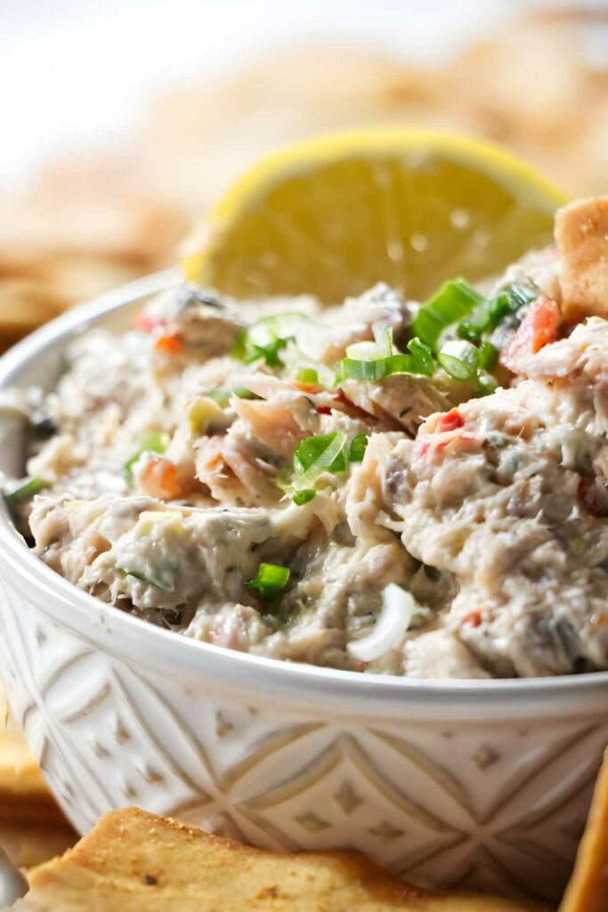 Close-up of creamy salmon spread with green onions and lemon, pita chips in background.