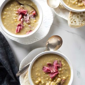 Three bowls of soup with shredded meat, corn, bread, and spoons on marble.