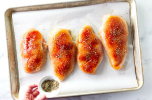 Adding herbs de Provence to chicken breasts on a sheet pan.
